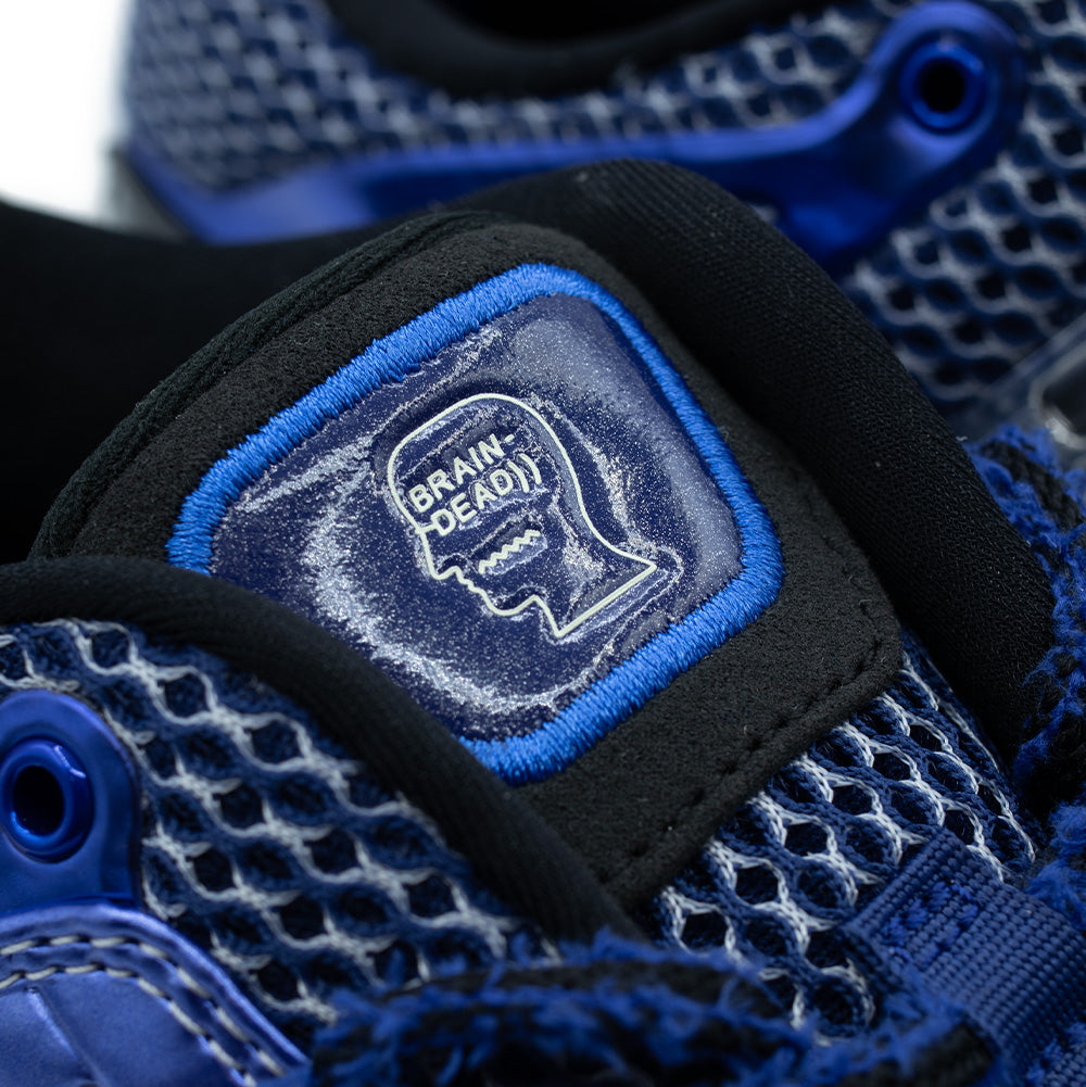 Macro close-up shot of the tongue badge on the adidas x Brain Dead Barricade V, highlighting the embossed white Brain Dead profile head logo inside a glossy dark blue badge bordered by electric blue satin embroidery, set over breathable open-cell mesh.