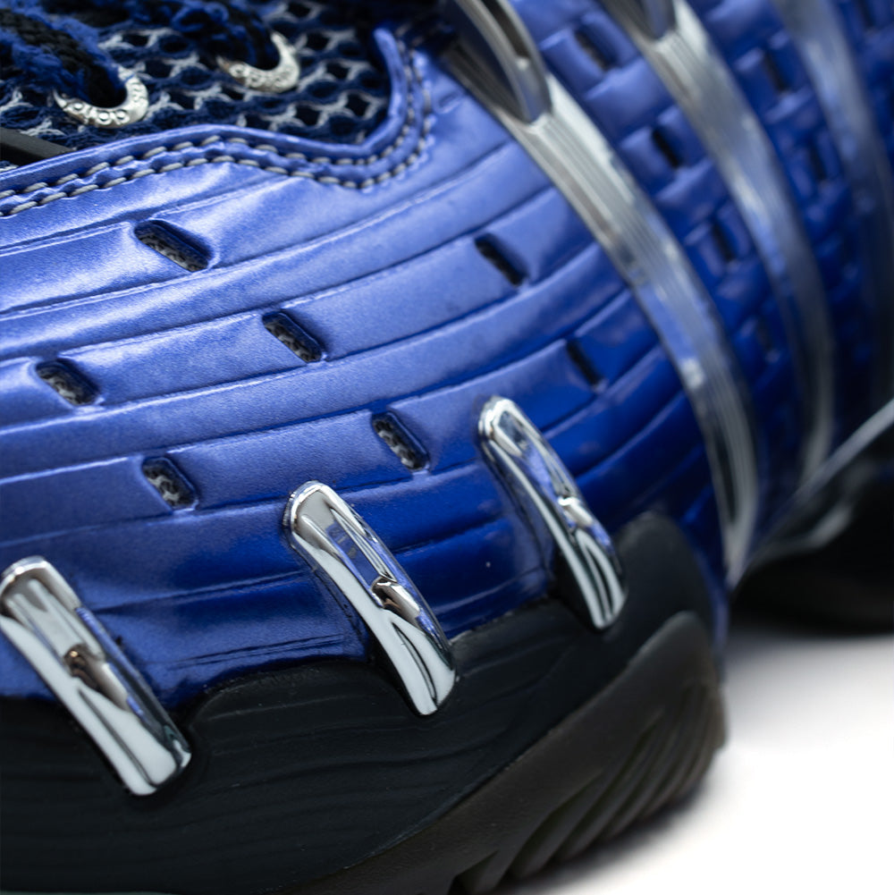 Extreme macro close-up of the lateral forefoot on the adidas x Brain Dead Barricade V, displaying the metallic Mystery Ink blue ribbed shell, ventilation cutouts, shiny chrome claw accents, and textured black lateral outsole wrap.