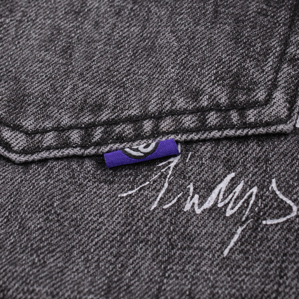 Studio macro detail of the rear pocket edge on the Always Waza Jeans, highlighting the vivid purple mini woven flag label displaying a white circular @ emblem, clean pocket seam topstitching, and white cursive graffiti lettering printed onto the faded black cotton denim.