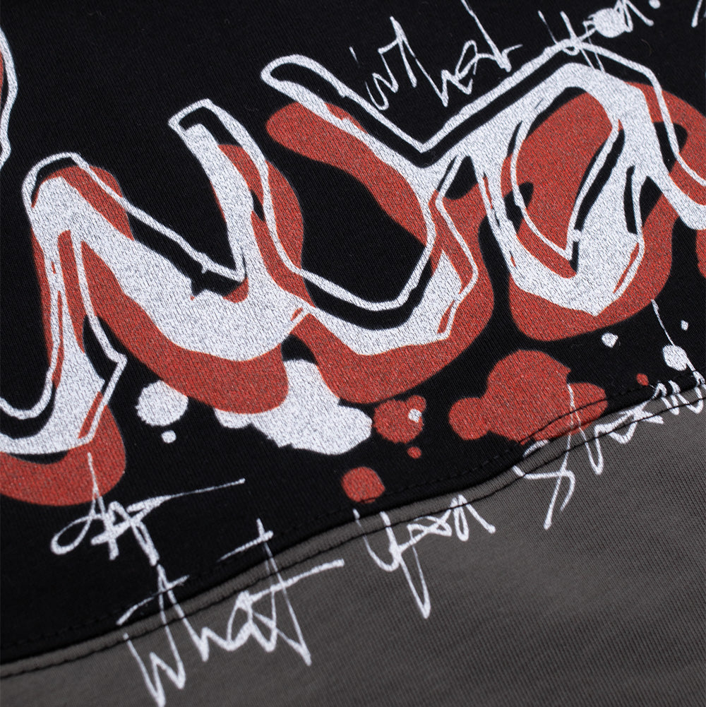 Detailed macro close-up of the front chest graphic on the Always Waza Panelled T-Shirt, highlighting the textured white screen-printed street-art lettering, bold red and orange spray texture, white scribble typography, and the clean horizontal seam connecting the black upper panel to the charcoal grey cotton jersey.