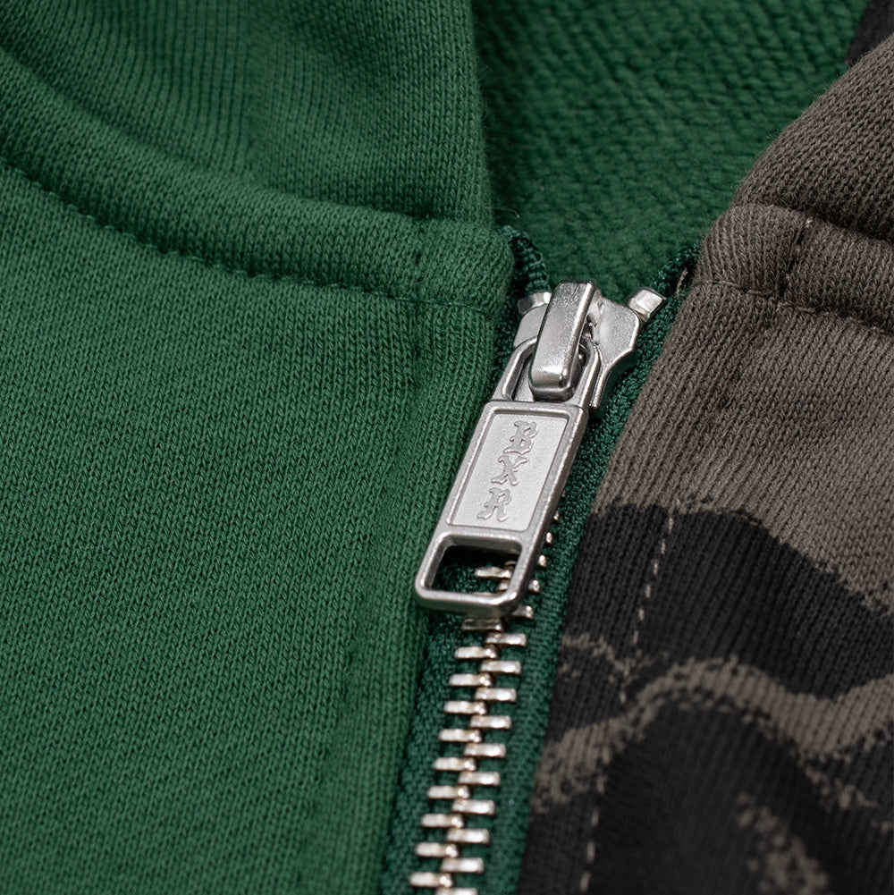 Detailed macro studio close-up of the metal front closure on the Born X Raised Mashup Hoodie, displaying the polished silver metal zipper track and branded rectangular puller engraved with the Old English Gothic "BXR" logo, joined between the green and brown fleece panels.