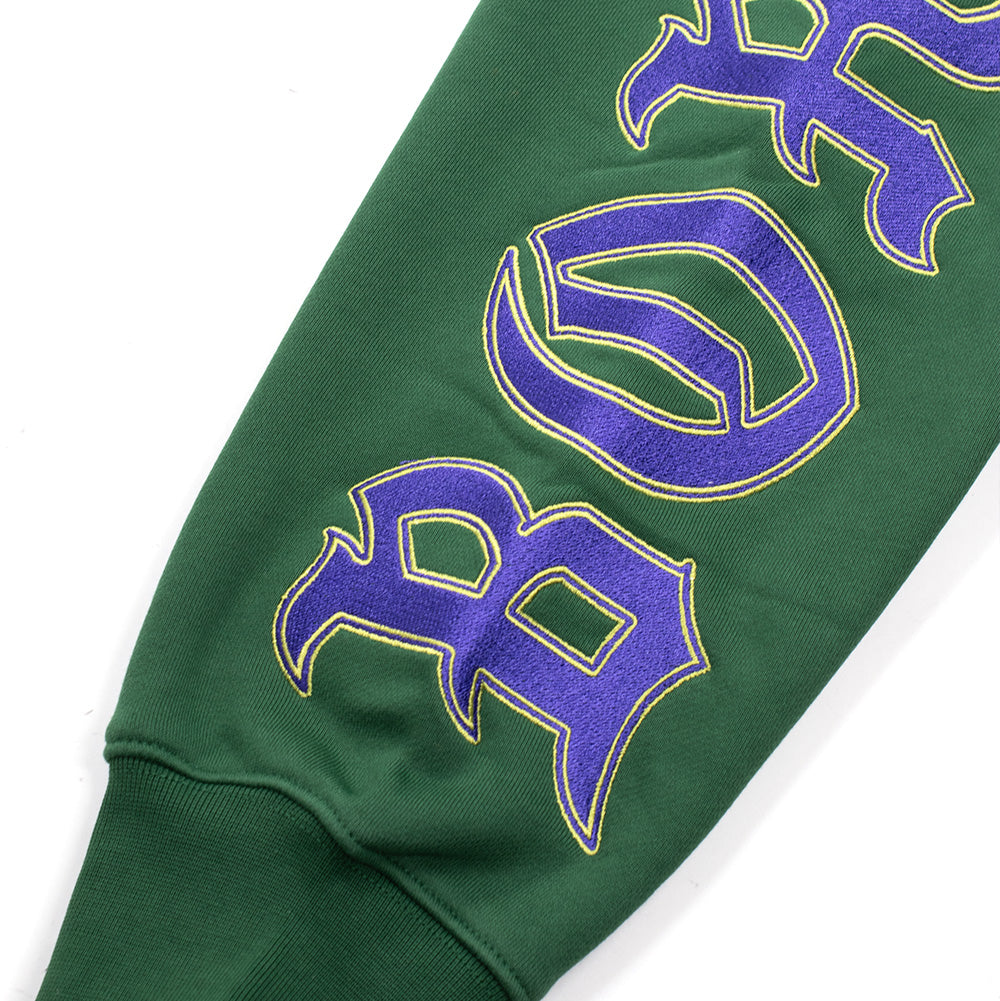 Detailed macro perspective of the sleeve graphics on the green arm of the Born X Raised Mashup Zip-Up Hoodie, highlighting the textured purple Old English Gothic letters outlined in vibrant yellow ink spelling out the brand name above the ribbed elastic cuff.