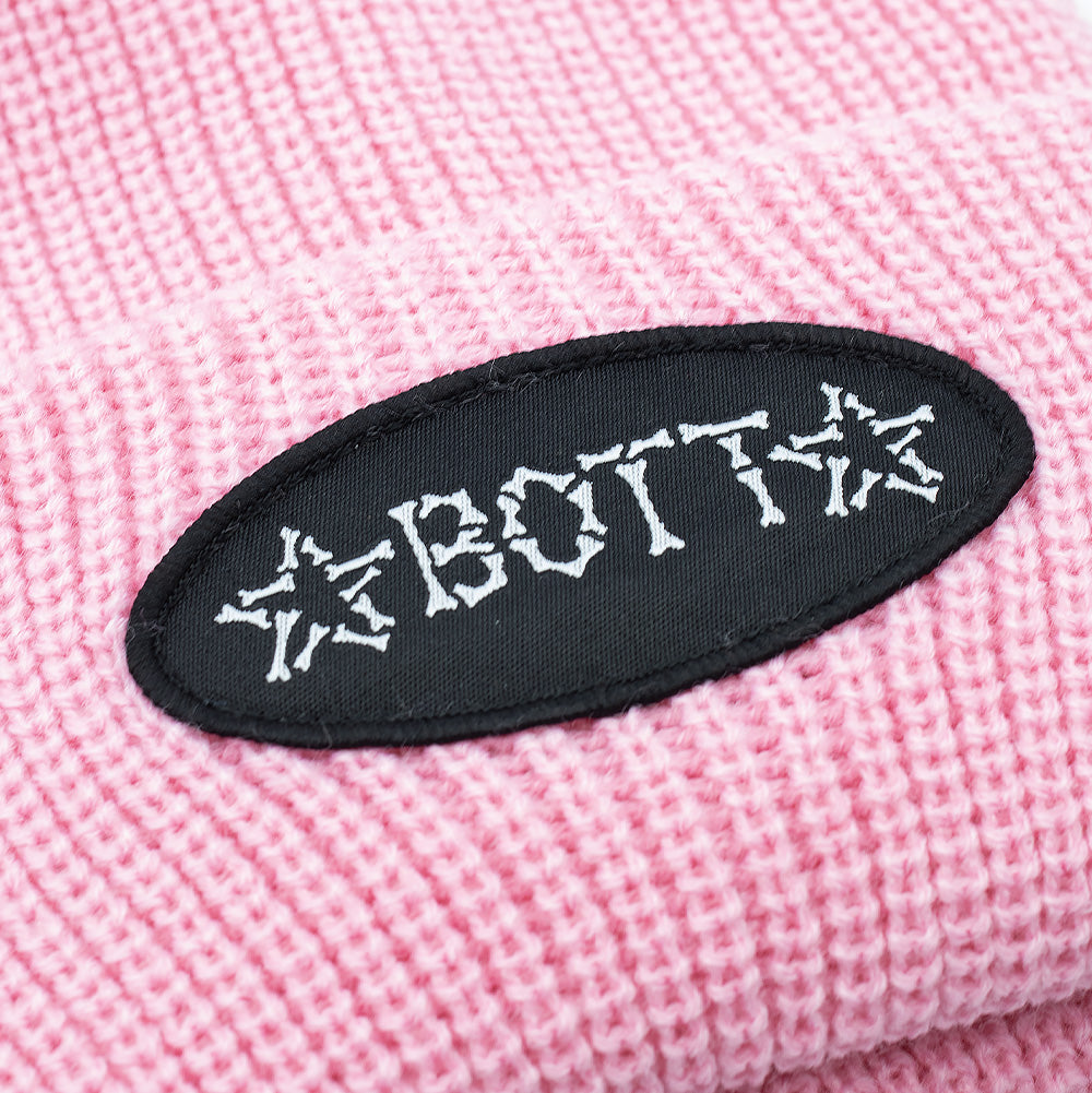 Detailed macro studio close-up of the folded cuff on the pink BoTT Bones Cuff Beanie, capturing the chunky elasticated rib-knit texture and the black oval fabric patch densely embroidered with white thread forming the bone-constructed "BoTT" logo flanked by matching bone stars.