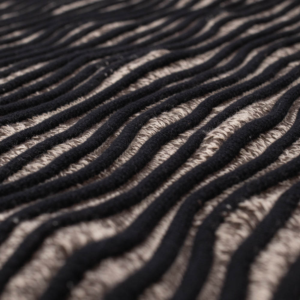 Extreme macro angled perspective of the knit surface on the Brain Dead 3D Wave Cropped Sweater, capturing the heavy, tactile depth of the raised serpentine black rope-like piping flowing horizontally over the heathered, open-weave ash taupe cotton yarn beneath.