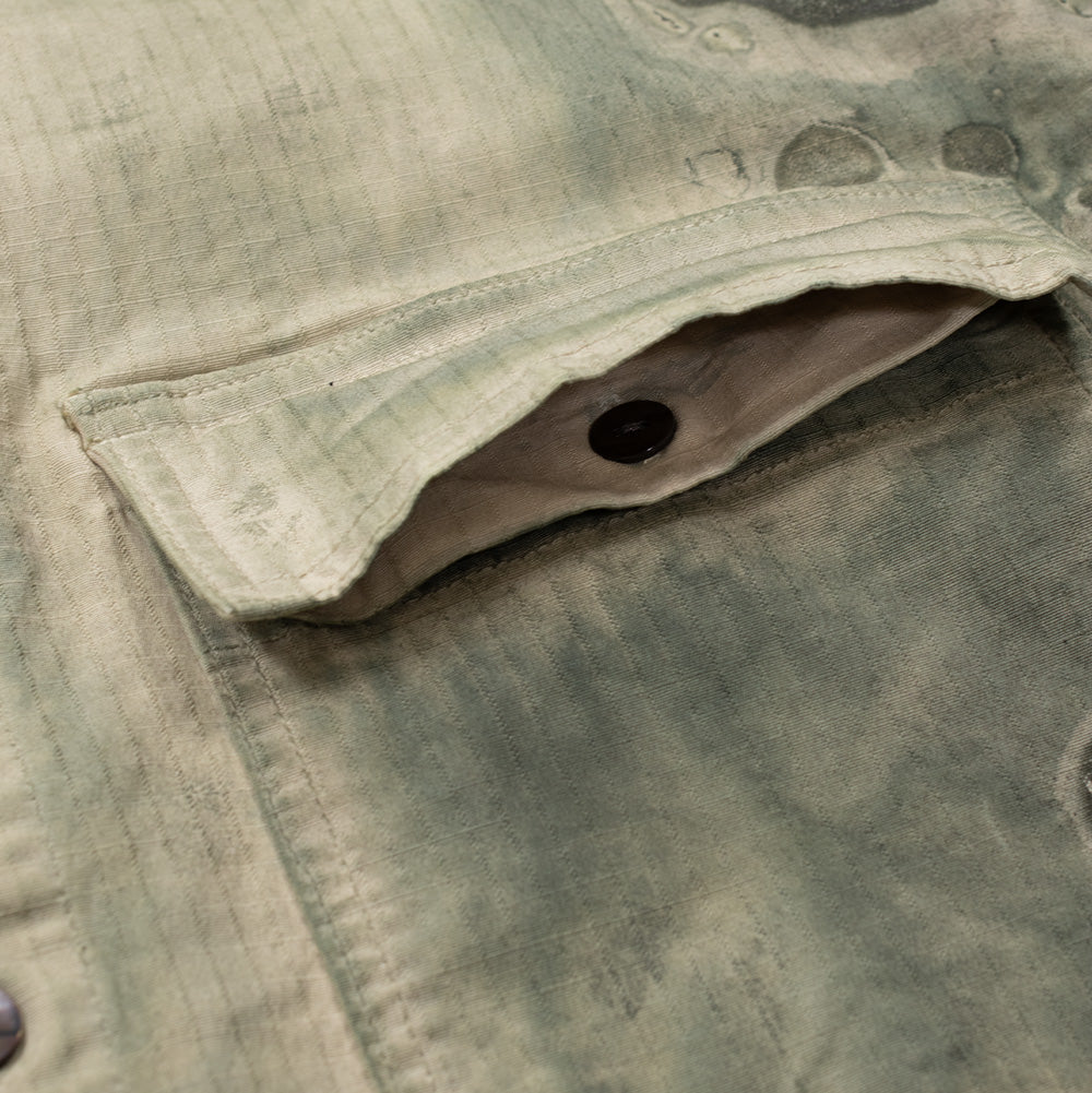 Macro studio view of the right chest flap pocket on the Brain Dead Cotton Ripstop Shirt Jacket, displaying the concealed button-down closure underneath the flap, durable double-needle stitch detailing, and the mottled greenish-grey organic overprint covering the pocket facade.