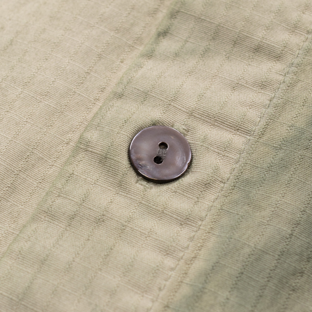 Extreme macro close-up of a dark genuine mother-of-pearl button stitched onto the placket of the Brain Dead Cotton Ripstop Cropped Shirt Jacket, highlighting its glossy, reflective mineral sheen against the fine grid structure of the pre-washed sand-colored ripstop textile.