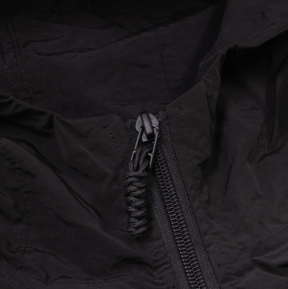 Detailed macro studio close-up of the neckline and hood on the Brain Dead Dolman Sleeve Jacket, showcasing the matte black metal two-way zipper puller with a thick braided climbing-style paracord tab, sturdy reverse-coil zipper track, and clean double-stitched collar seams on the textured black nylon fabric.
