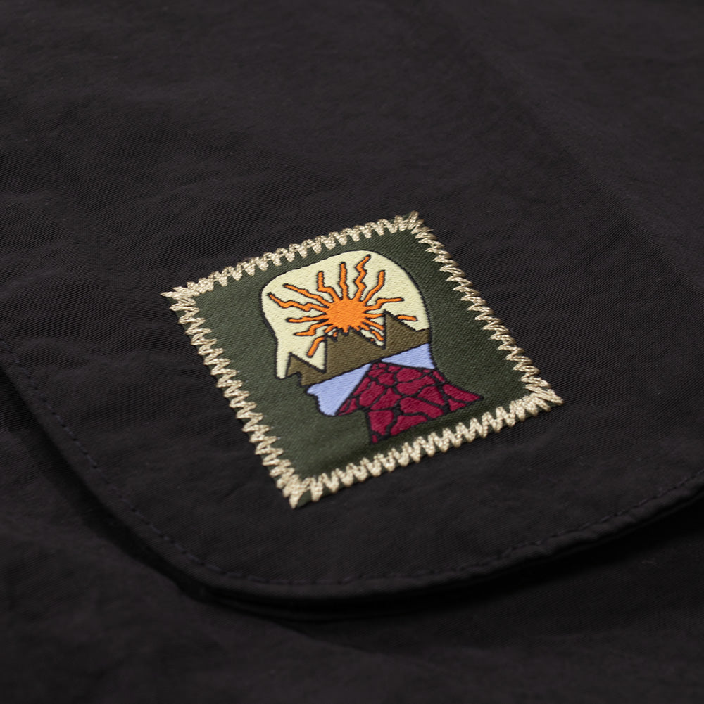 Detailed close-up shot of the woven brand patch on the lower left front pocket of the Brain Dead Dolman Sleeve Jacket, displaying the colorful embroidered silhouette of the Brain Dead head filled with a rising sun behind mountain peaks, framed by olive-drab fabric and secured by heavy cream zigzag edge stitching.