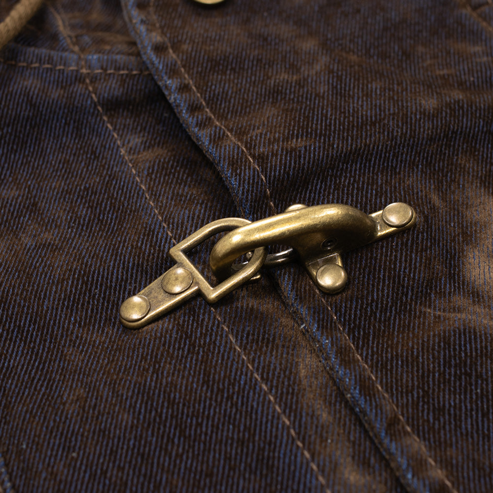 Macro studio view of the signature hardware on the Brain Dead Flocked Denim Fireman Jacket, focusing on a heavy antique brass metal fireman clip clasp securely hooked into a riveted D-ring fastener across the central front placket.