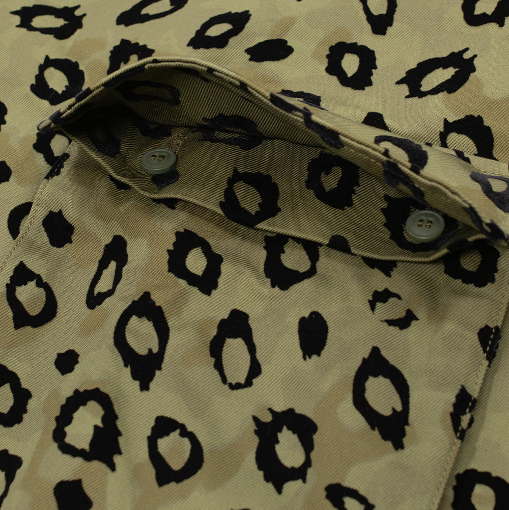 Macro studio view of the right lower flap pocket on the Brain Dead Hooded Chore Coat, showing the opened flap revealing hidden dual olive-green interior buttons and reinforced buttonholes, framed by thick three-dimensional black flocked leopard spots.