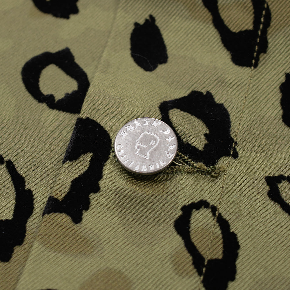 Close-up shot of the front placket closure on the Brain Dead Leopard Camo Coat, focusing on a branded metallic snap button featuring the signature etched Brain Dead profile emblem, surrounded by raised, soft-touch black flocking on textured khaki-green cotton canvas.
