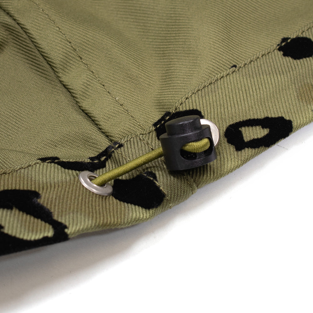 Macro studio perspective of the bottom hem cinch system on the Brain Dead Hooded Chore Coat, capturing the silver-tone metal grommet, olive green elastic bungee shock cord, and adjustable black spring toggle stopper set against the folded interior hem facing.
