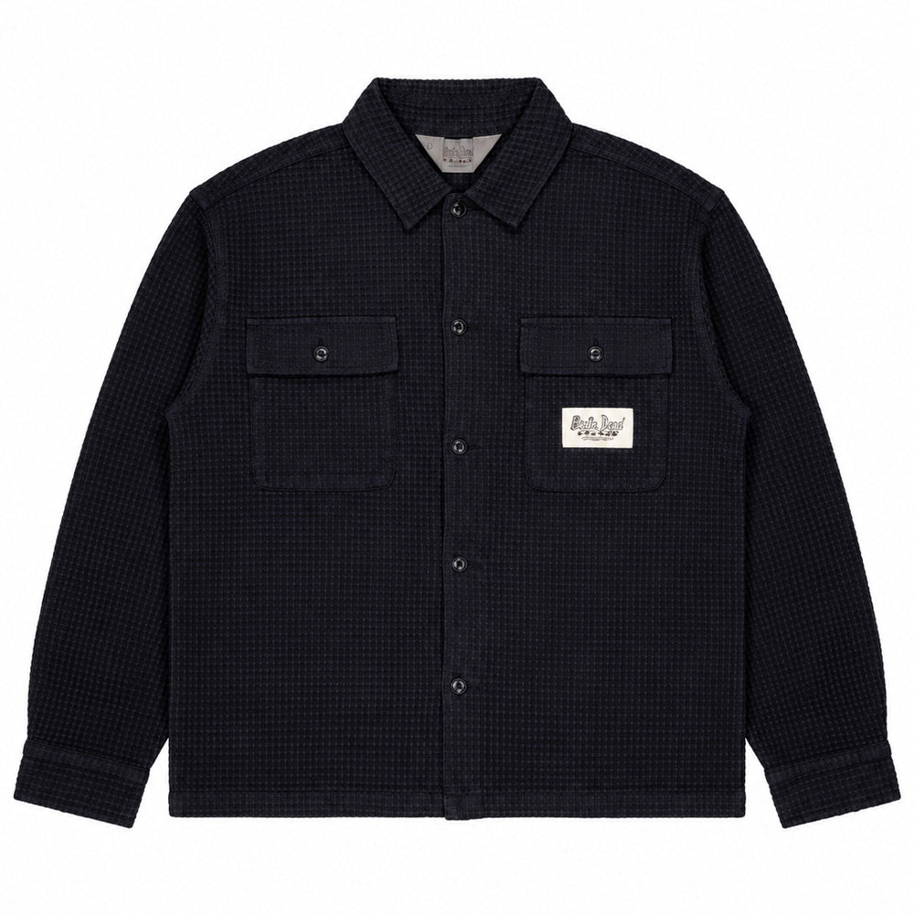 Front flat lay studio view of the Brain Dead Waffle Button Front Shirt in washed black, showcasing a boxy workwear-inspired overshirt silhouette crafted from heavy waffle-textured cotton weave, featuring a classic spread collar, black button closure placket, two symmetrical buttoned flap chest pockets, straight hem, and a white woven rectangular Brain Dead California Designs label stitched onto the left pocket.