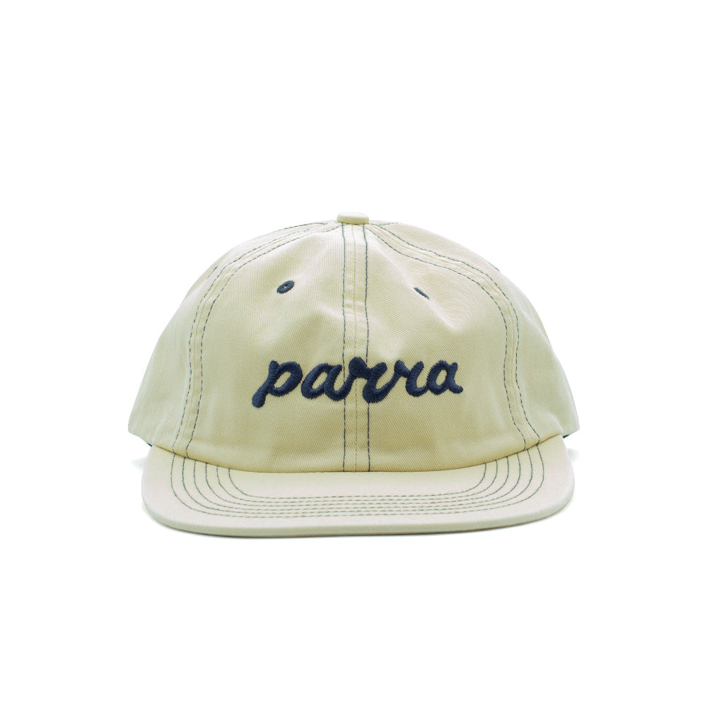 Front perspective studio view of the By Parra Lowercase Script Logo 6 Panel Hat in natural off-white pre-washed cotton twill, showcasing an unstructured low-profile polo silhouette, curved visor with multi-row contrast stitching, dark navy embroidered ventilation eyelets, and navy blue chain-stitched cursive lowercase "parra" lettering across the front crown.
