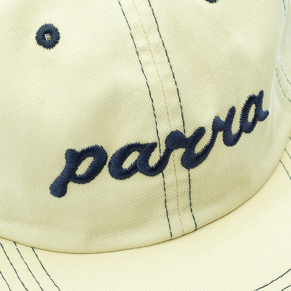 Detailed macro studio close-up of the front crown on the By Parra 6 Panel Hat in Natural, capturing the thick, raised dark navy blue embroidery forming the flowing lowercase cursive "parra" logo against the diagonal texture of the washed cream cotton twill fabric.