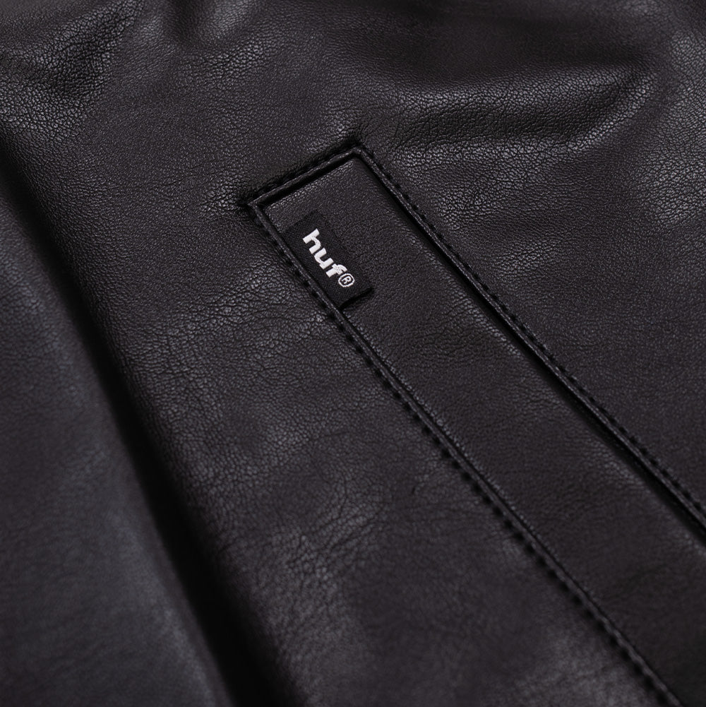 Macro view of the front welt pocket on the black Huf X Three 6 Mafia jacket, focusing on the stitched pocket reinforcement and the woven black HUF brand flag label sewn onto the upper welt seam.