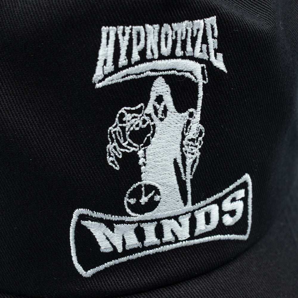 Detailed macro studio close-up of the front panel on the HUF x Three 6 Mafia Snapback, highlighting the intricate white thread embroidery of the Hypnotize Minds record label graphic showing the hooded Grim Reaper figure, hourglass, scythe, and framed banners against black woven twill fabric.