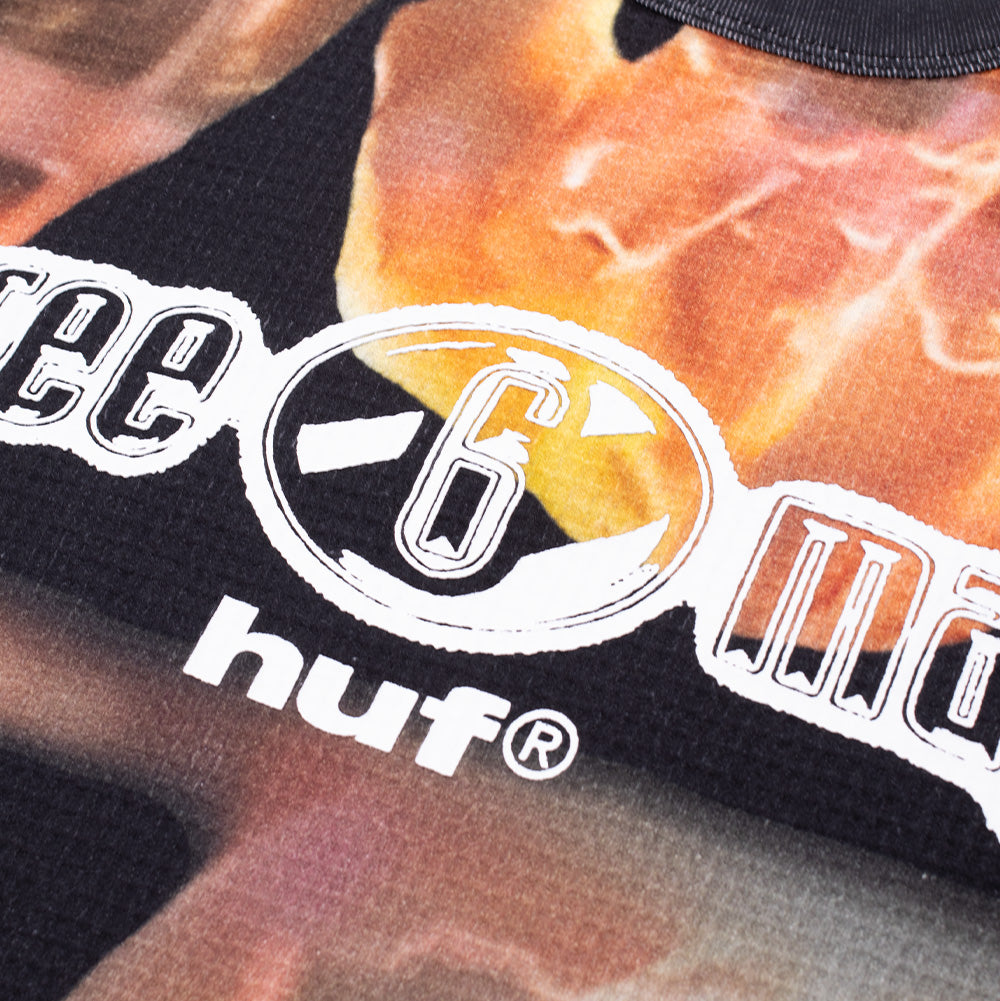 Detailed macro studio close-up of the front chest graphic on the Huf X Three 6 Mafia Torched Longsleeve Thermal, highlighting the white-outlined chrome "three 6 mafia" lettering with an orange-tinted circular "6" badge, set against the fiery textured honeycomb thermal knit pattern.