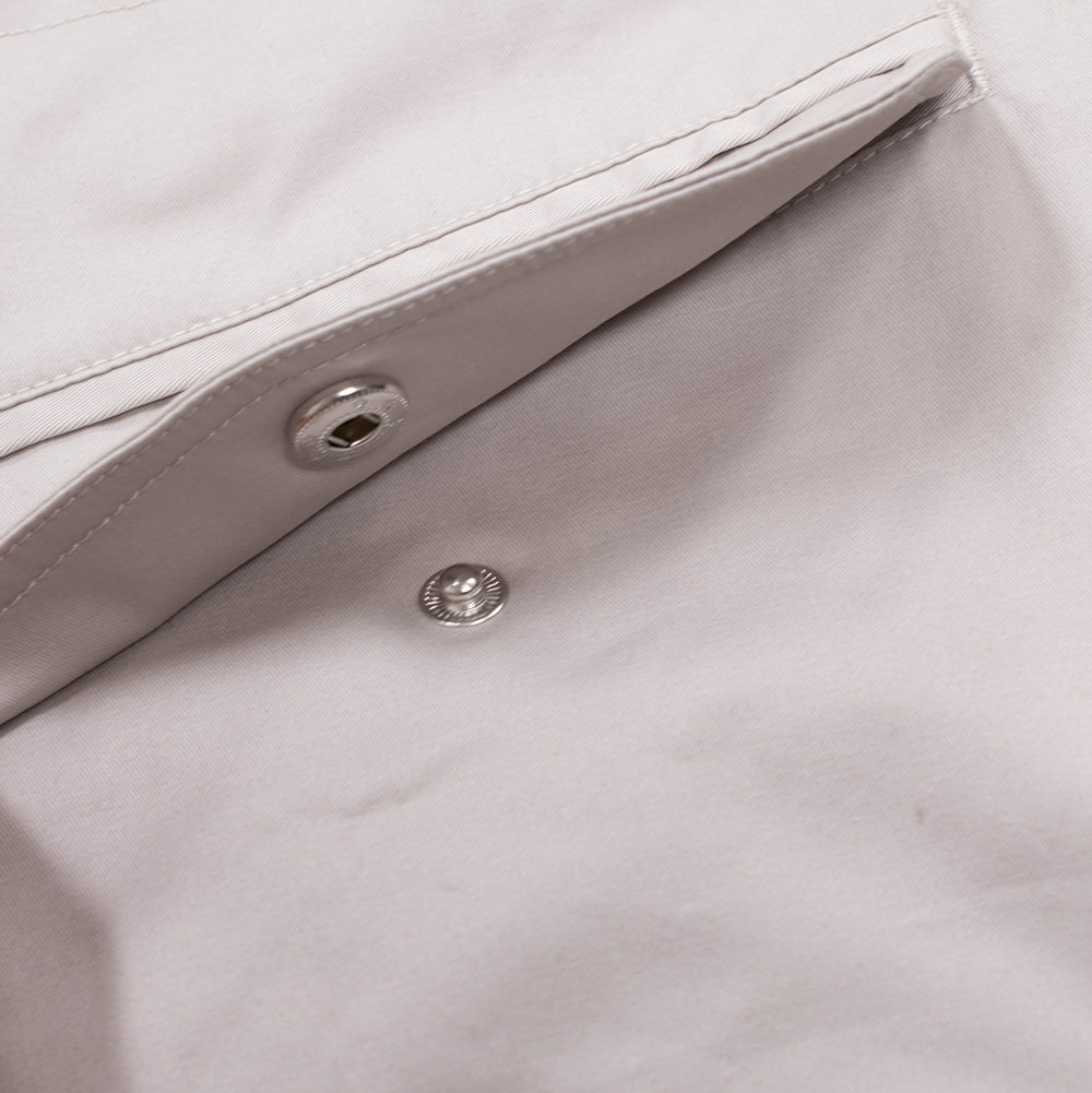 Macro studio view of the front pocket flap on the Metalwood Studio Cotton Twill Parka, slightly parted to show the glossy silver metallic snap-button fastener and clean tonal double topstitching on the stone twill fabric.