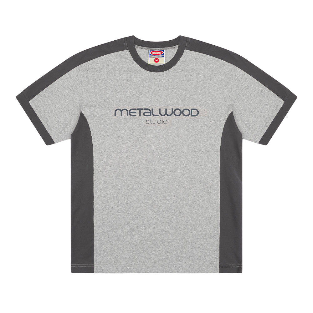 Front flat lay studio view of the Metalwood Studio Creature T-Shirt in Heather Grey, showcasing a boxy streetwear cut with contrasting dark charcoal grey curved shoulder yokes and side body panels, a charcoal rib-knit crewneck collar and sleeve cuffs, and centered blue-grey "METALWOOD studio" logo lettering printed across the chest.