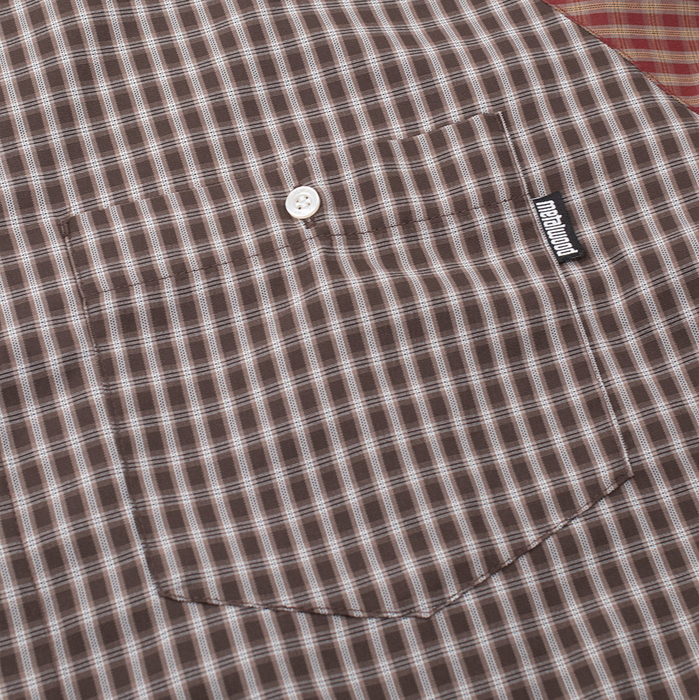 Detailed macro perspective of the chest pocket on the Metalwood Studio Frankenstein Button Up Shirt, highlighting the brown plaid fabric pattern alignment, centered white pearlescent button closure, red raglan shoulder seam edge, and vertical black woven "metalwood" flag label.