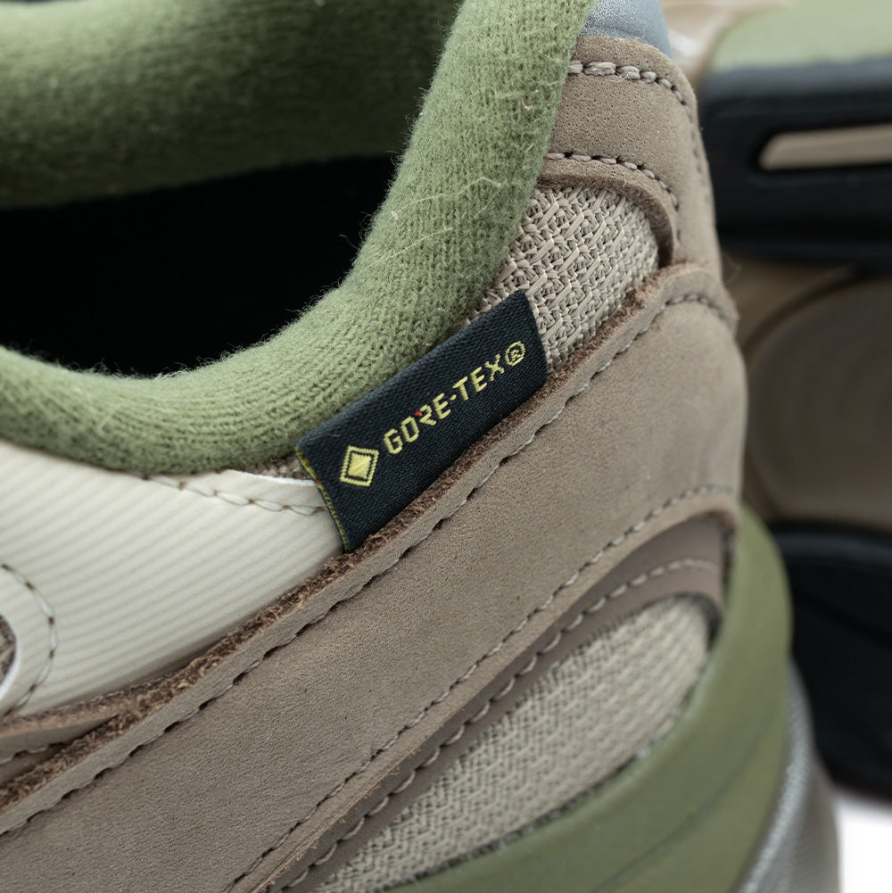 Extreme macro close-up of the lateral ankle collar on the New Balance 2010 GORE-TEX, highlighting the black woven technical GORE-TEX flag tag with gold lettering stitched beside the plush sage green inner fleece lining and premium tan nubuck leather overlays.
