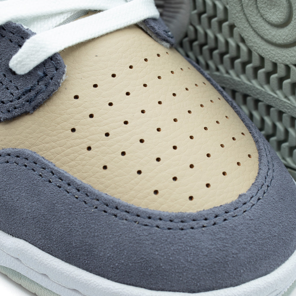 Detailed macro studio close-up of the toe box on the New Balance Numeric 480, displaying the fine-textured pebble grain of the incense tan leather toe panel with ventilation perforations, bounded by double-stitched meteorite blue-grey suede ollie guards.