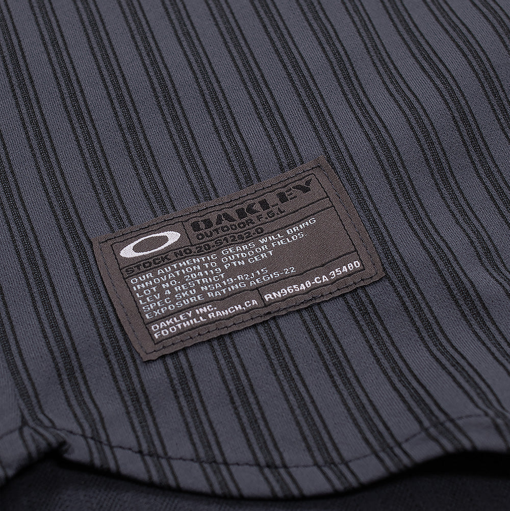 Macro studio view of the lower hem branding on the Oakley FGL Fuel LS Shirt, focusing on the dark woven military-style spec patch detailed with the white Oakley Ellipse icon, "OAKLEY OUTDOOR F.G.L." lettering, and authentic product codes against the vertically striped fabric.