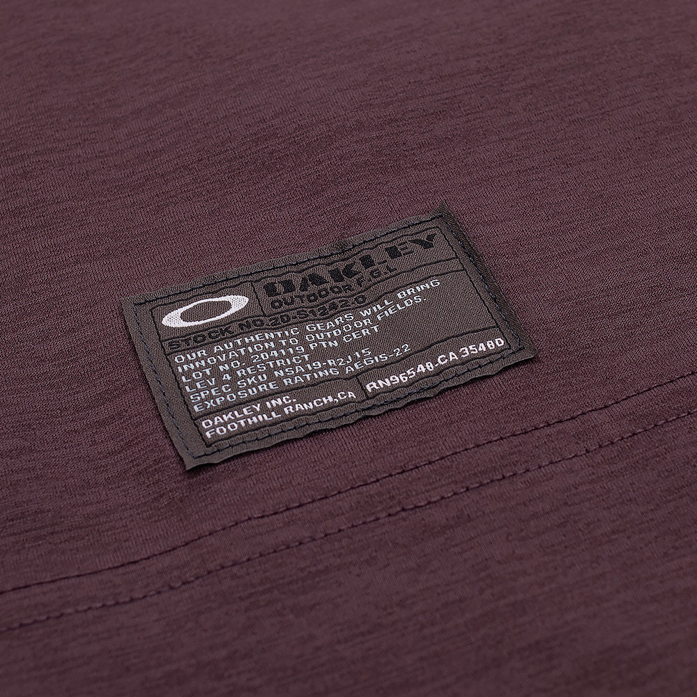 Macro studio close-up of the hem label on the Oakley FGL Scratch SS Tee 6.7 in Rouge, showcasing the rectangular dark grey woven utility tag with white Oakley Ellipse branding, "OAKLEY OUTDOOR F.G.L." lettering, and field issue specification codes stitched securely above the bottom hem.