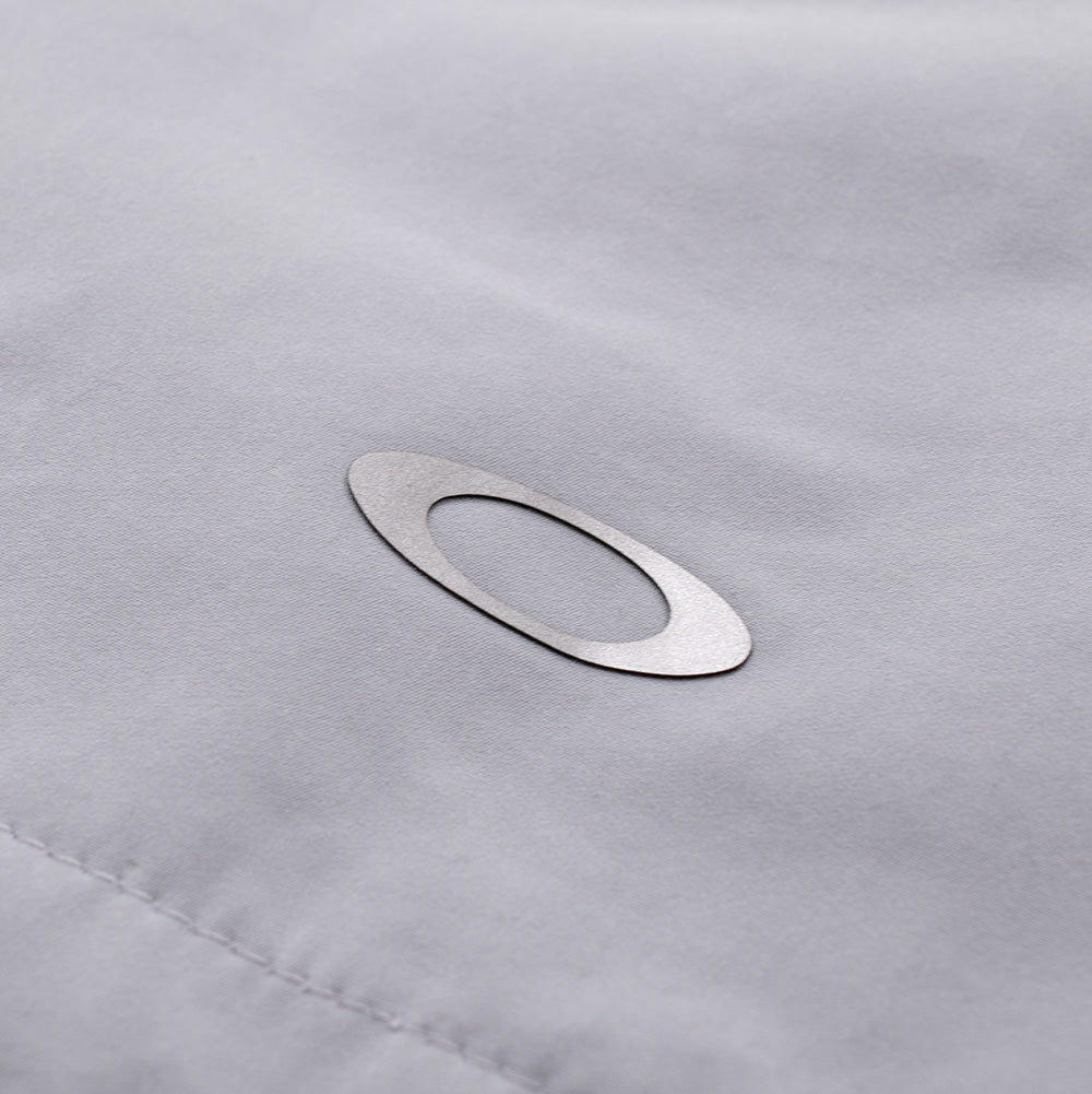 Extreme macro close-up of the rear hem detailing on the Oakley FGL Sector Jacket, displaying the smooth, powdery texture of the water-repellent Winter Grey hybrid weave accented by a precision-bonded reflective silver metallic Oakley Ellipse logo.