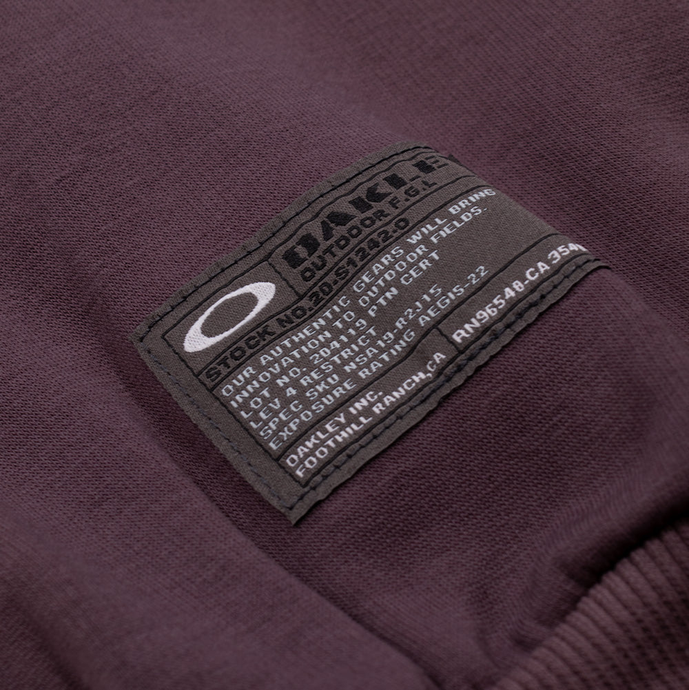 Macro studio view of the sleeve cuff branding on the Oakley FGL Slick Fleece Hoodie 6.7 in Rouge, showcasing the rectangular charcoal military-spec woven patch with white Oakley Ellipse branding and outdoor technical specifications stitched above the ribbed wrist cuff.
