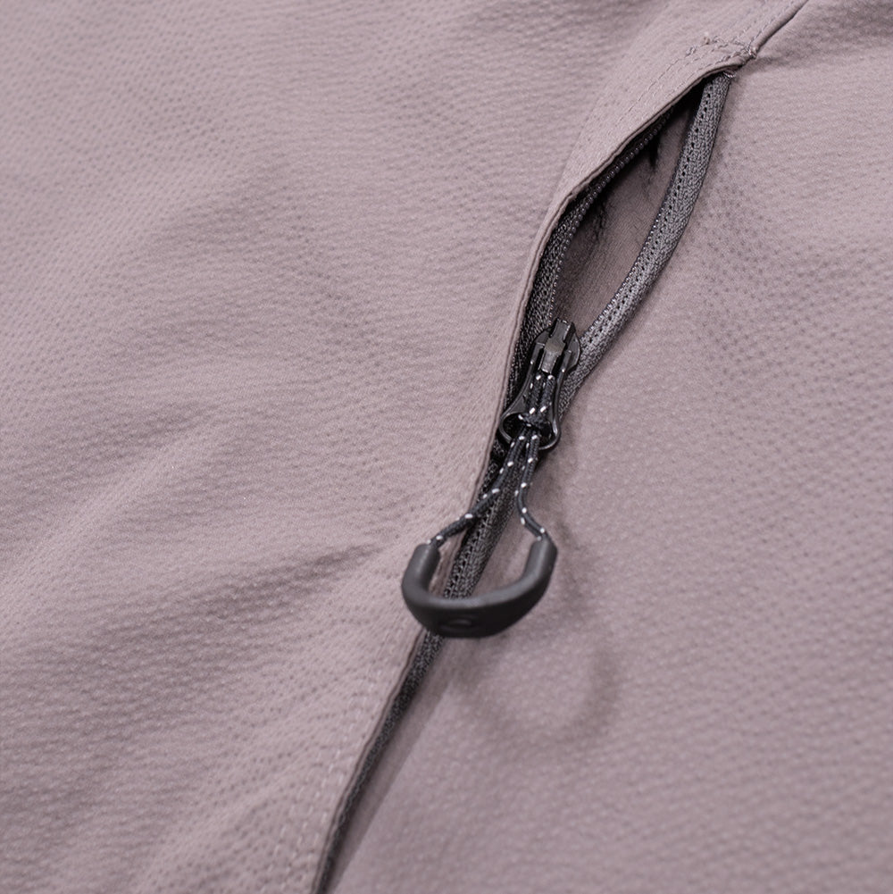 Macro close-up shot of the lower hand pocket opening on the Oakley FGL Stroke Anorak, detailing the concealed vertical zipper track, reinforced seam stitching, and a black reflective-flecked paracord zipper pull loop with a curved ergonomic rubber grip set against the textured Gray Wash fabric.