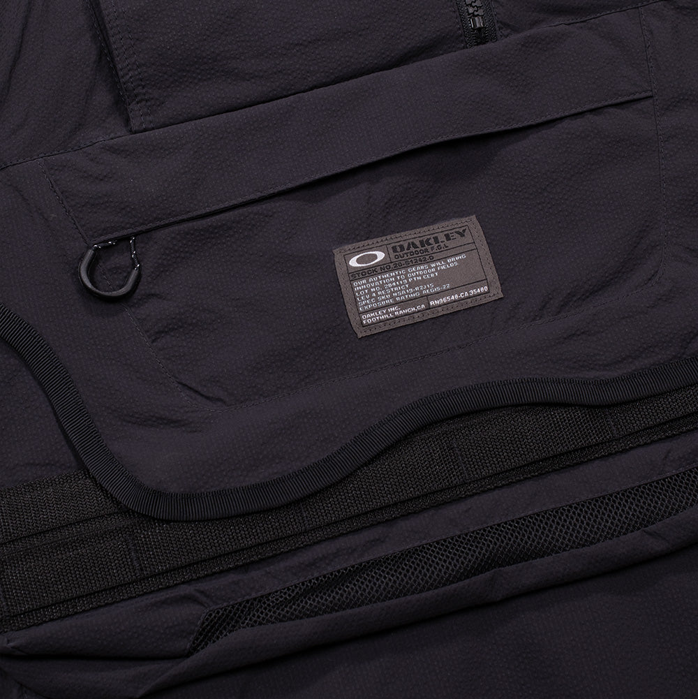 Macro studio view of the front chest utility pocket on the Oakley FGL Stroke Anorak, showing the curved protective flap, horizontal zipper pocket with cord loop puller, dual tactical webbing gear loops, breathable bottom mesh trim, and a dark rectangular woven military-spec patch displaying the white Oakley Ellipse logo and technical specification codes.