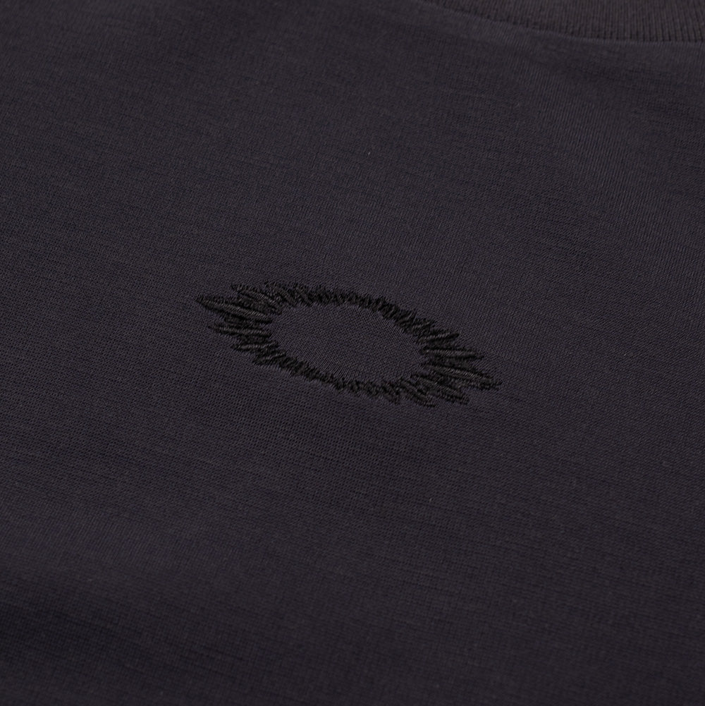 Macro studio close-up of the front chest embroidery on the Oakley FGL Union SS Tee Phantom, highlighting the tonal black embroidery thread creating a jagged, radiated contour around the Oakley Ellipse icon against the dark charcoal technical performance jersey.