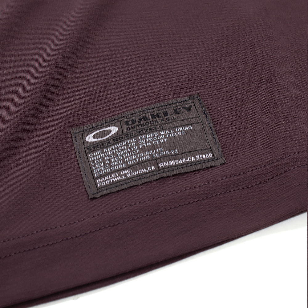 Macro studio view of the lower hem branding on the Oakley FGL Union SS Tee 6.7 in Rouge, showing the charcoal military-specification woven label with the white Oakley Ellipse logo, "OAKLEY OUTDOOR F.G.L." text, and technical product ratings stitched above the double-needle bottom seam.