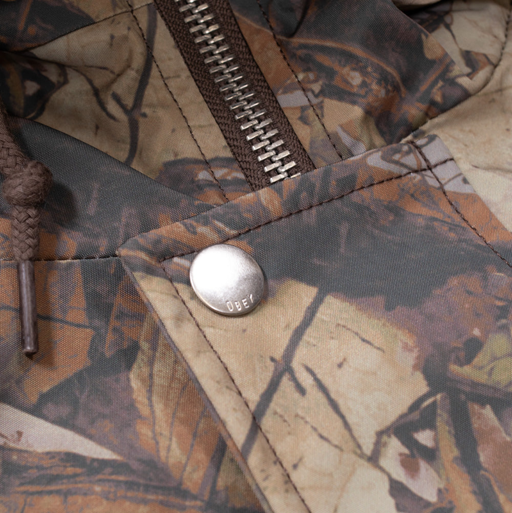 Macro studio close-up of the upper collar closure on the Obey N-2B Short Parka, detailing the chunky antique silver metal zipper track, woven brown drawstring cord, and a brushed silver metal snap button engraved with subtle OBEY branding on the camo storm placket.