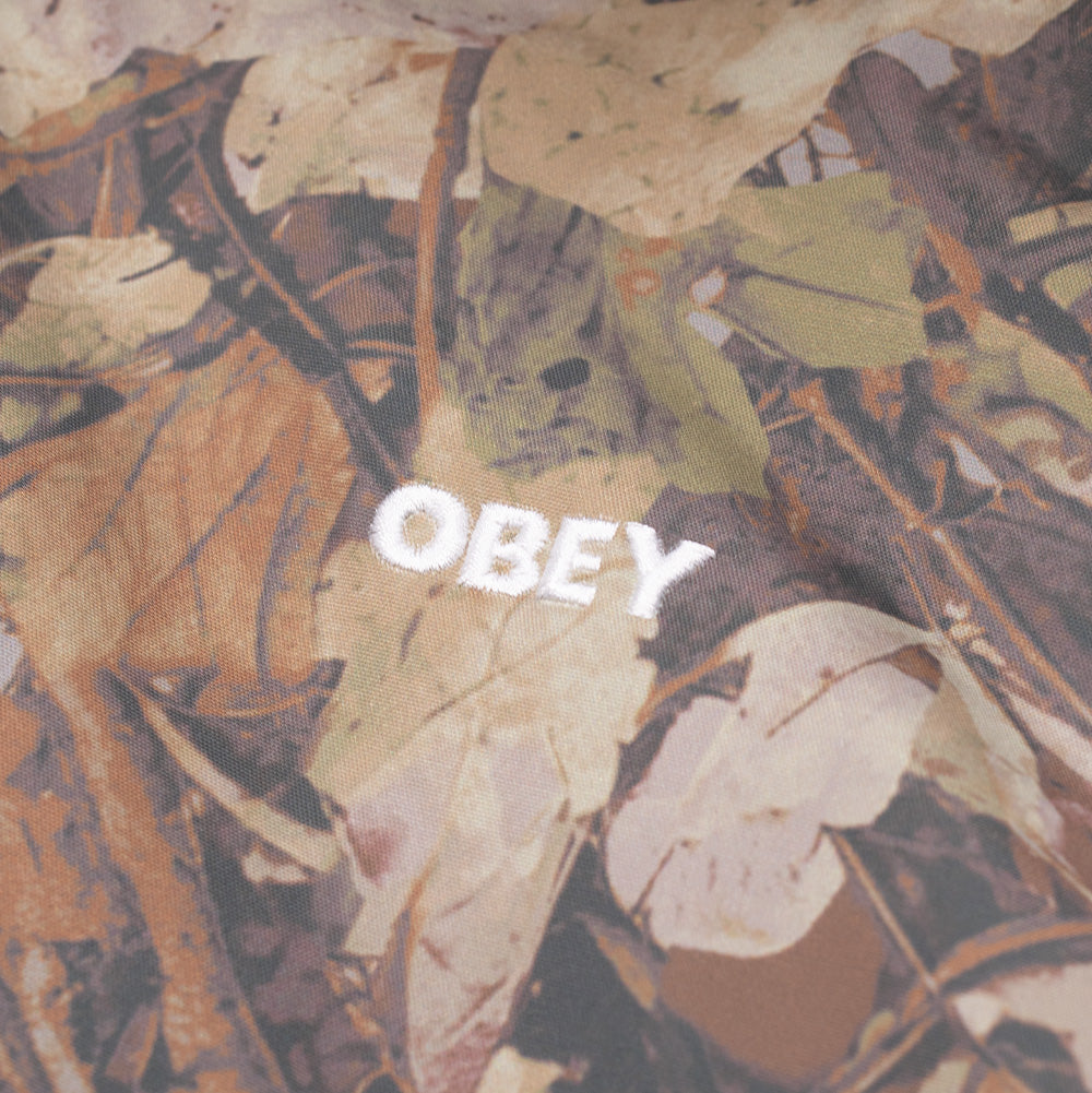 Detailed close-up shot of the left chest branding on the Obey N-2B Short Parka, highlighting the clean, dense white satin-stitched OBEY wordmark embroidery set against the printed autumn leaves and branch camo fabric.
