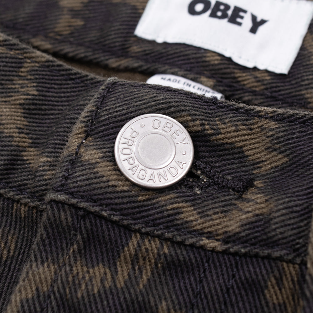Macro studio close-up of the waistband closure on the Obey Studded Boxcar Denim in Dark Leopard, detailing the silver metallic shank button embossed with "OBEY PROPAGANDA" lettering, dense diagonal denim twill weave in dark olive and black, and the woven white OBEY brand label inside the waistband.