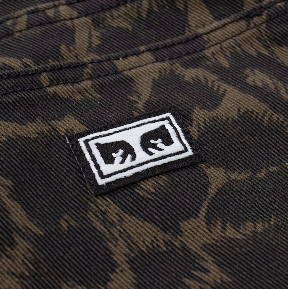 Studio macro perspective of the back right pocket on the Obey Studded Boxcar Denim, highlighting the miniature black woven rectangular tag embroidered with the signature white Obey Eyes Icon graphic, set against the dark textured leopard pattern denim fabric.