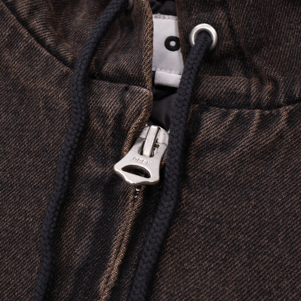 Macro studio close-up of the front zipper and collar closure on the Obey Studded Zip Up Jacket, highlighting the heavy silver metallic zipper with an engraved OBEY pull, black woven hood drawstring cords through metal grommets, and the textured rust-washed black denim twill fabric.