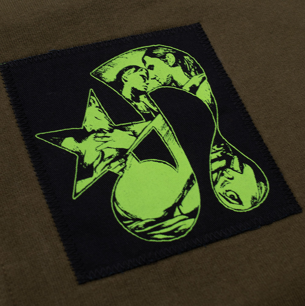 Close-up shot of the left chest patch on the Token Carly Zip Hoodie, featuring heavy zig-zag edge stitching around a black canvas patch printed with a vivid lime neon green artistic hourglass and kissing silhouettes with stars.