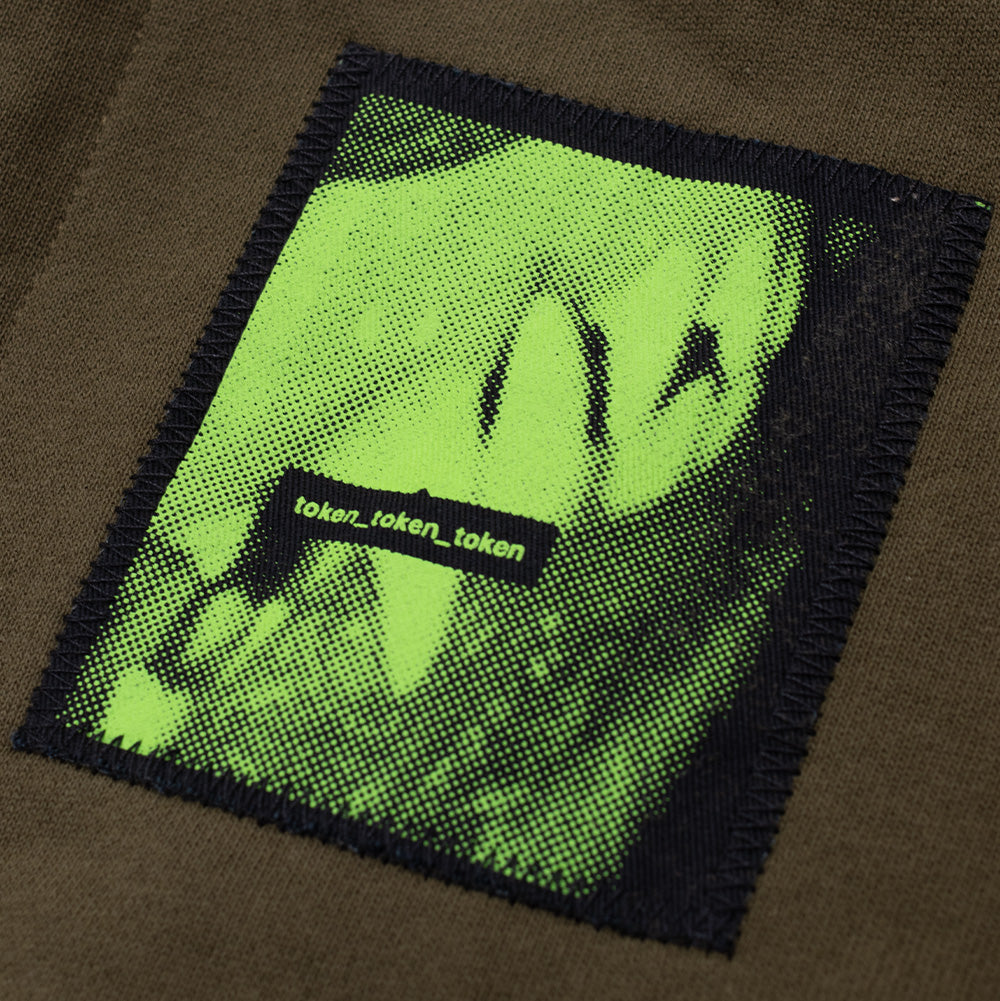 Studio macro shot of the top hood patch on the Token Carly Zip Hoodie, displaying heavy black perimeter stitching and a neon green halftone screen-printed graphic accented by a central black bar reading "token_token_token".