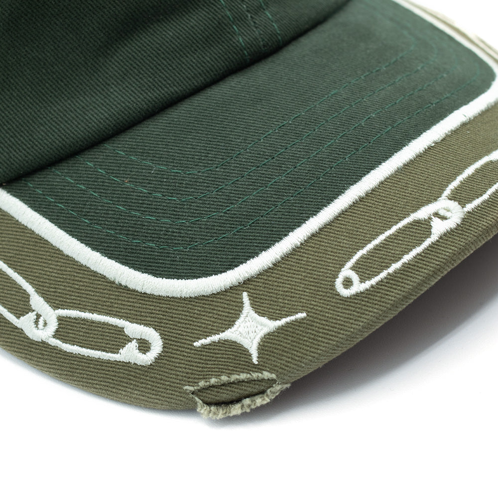 Macro studio view of the curved visor on the Token Lowkey Cap, detailing the contrast between the dark midnight green upper crown and the olive drab brim, framed by white embroidered looped safety pins, shining stars, curved line borders, and a frayed cut-out distressing along the visor edge.