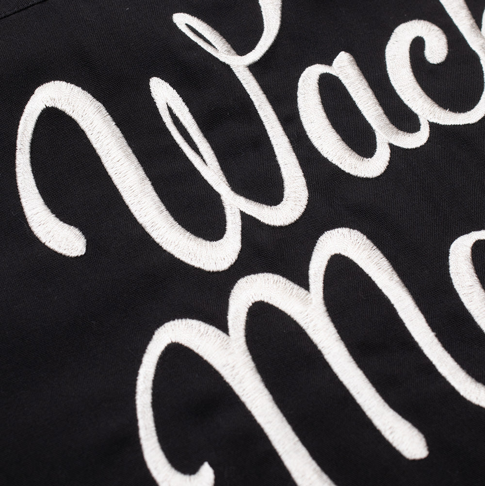 Extreme macro close-up of the back embroidery on the Wacko Maria 50's Shirt Type 5, focusing on the textured white satin rope stitching defining the flowing cursive letters of the "Wacko Maria" script against smooth, dark charcoal woven fabric.